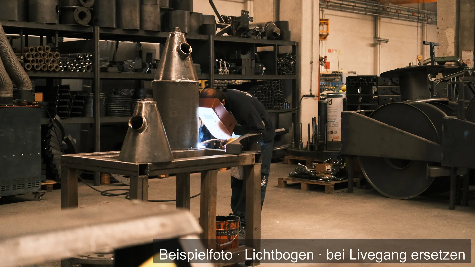 Werkstatt mit Stahlteilen und Maschinen; im Hintergrund schweißt ein Handwerker.