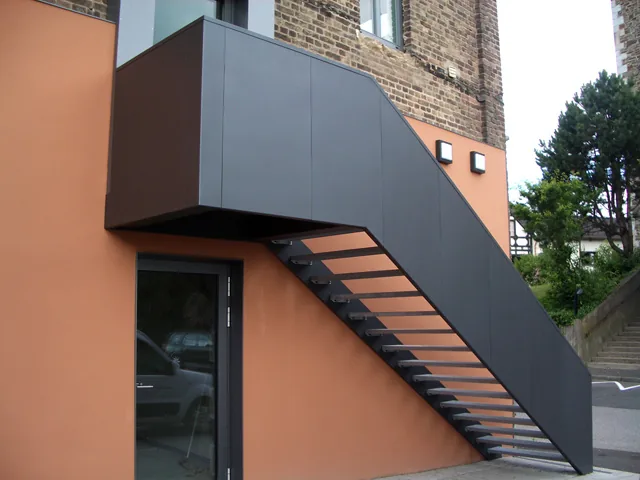 Außentreppe mit verkleideter Blechwange und offenen Stufen an einer orange verputzten Fassade.