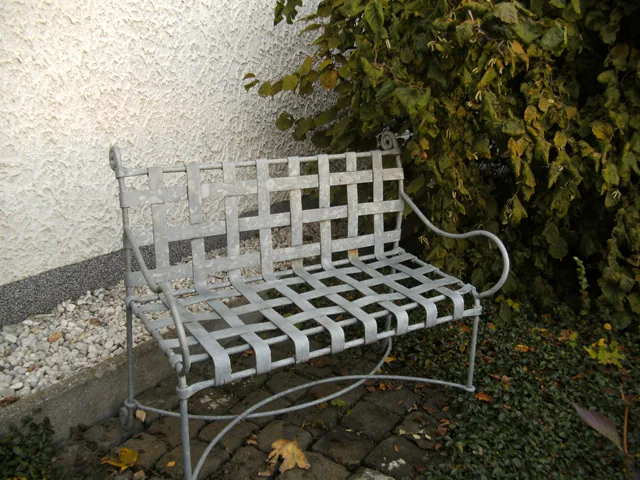 Gartenbank aus geflochtenem Flachstahl, verzinkt, vor einer Hauswand.