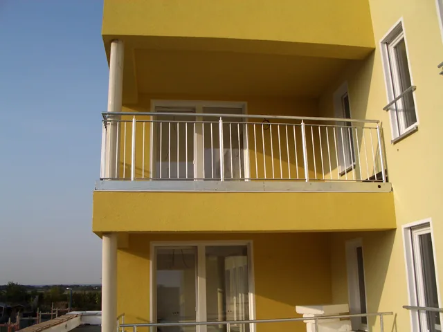 Balkon mit Stabgeländer aus Stahl an einer gelben Fassade.