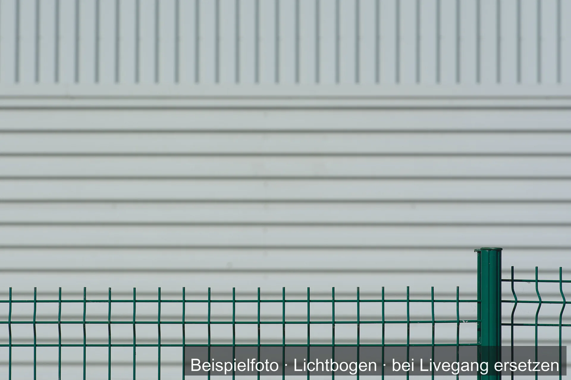Grüner Stabgitterzaun vor einer hellen Wand.